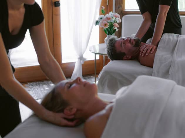 A serene spa setting featuring a couple receiving relaxing massages side by side, with natural light streaming through white curtains and a vase of flowers in the background