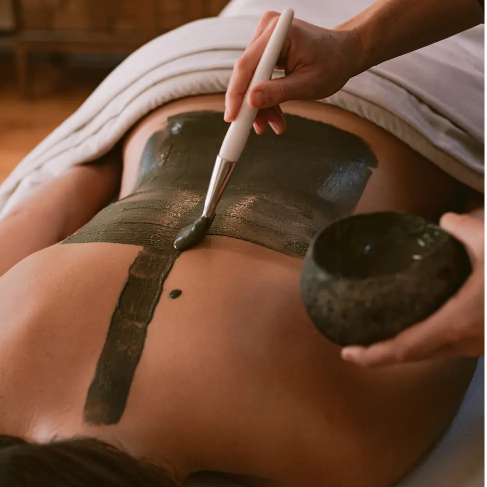 A person lying face down on a massage table with a towel covering the lower body while a practitioner applies a green mud or clay treatment to their back using a soft brush. The scene conveys relaxation and spa rejuvenation.