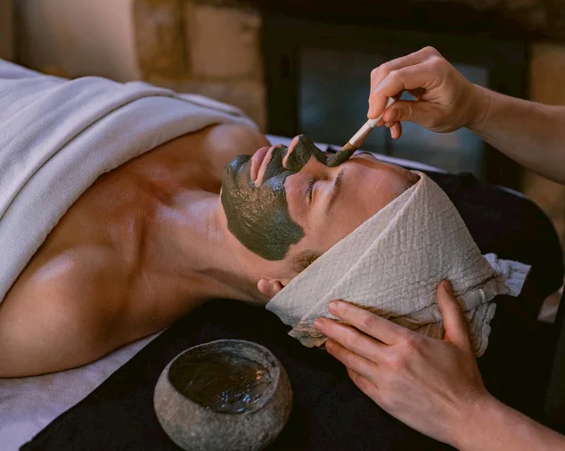 A client receiving a soothing facial treatment with a brush applying a dark mask, while their head is wrapped in a soft towel, set in a serene spa environment with a glowing fireplace in the background.
