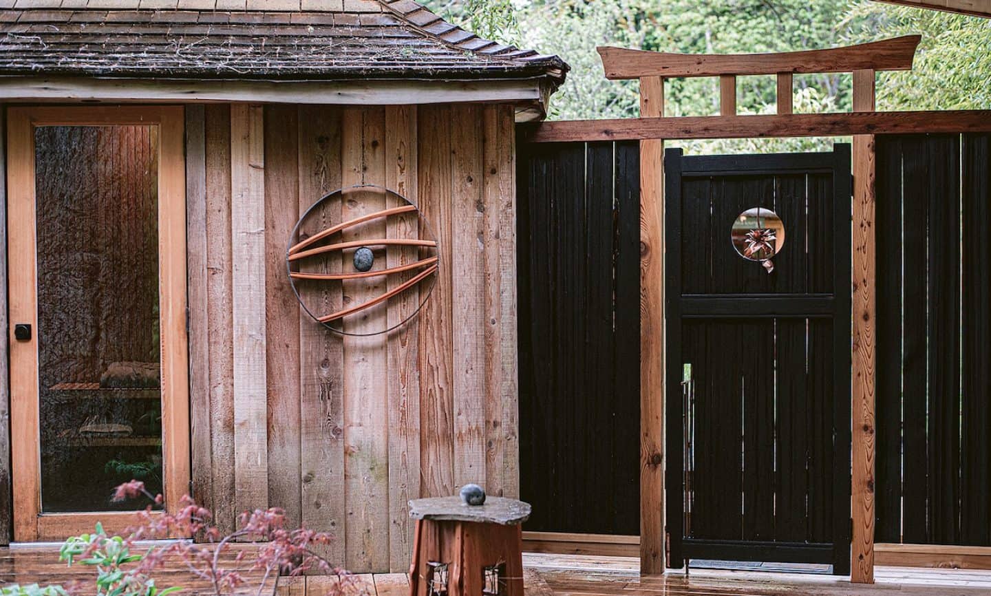 A tranquil outdoor setting featuring a rustic wooden sauna with a glass door, a handcrafted black wooden gate with a circular window, and natural decor elements. The peaceful environment is surrounded by lush greenery, creating a serene retreat.